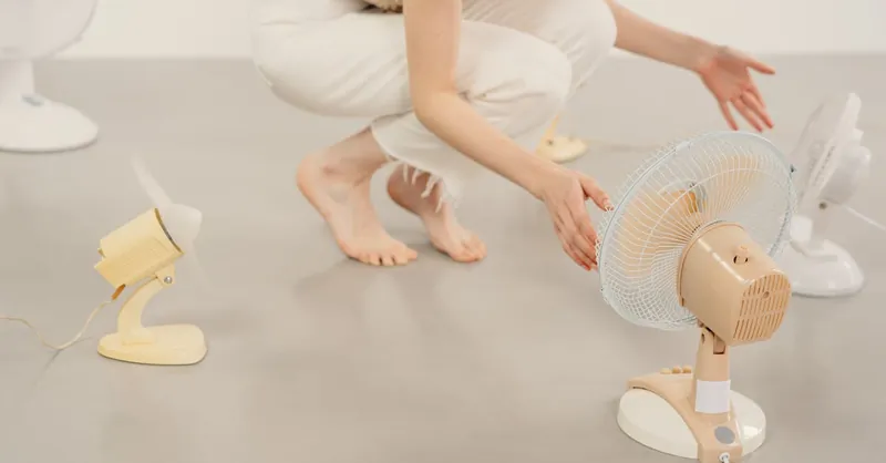 Barefoot person crouching to adjust multiple electric fans in a minimalist studio setting.
