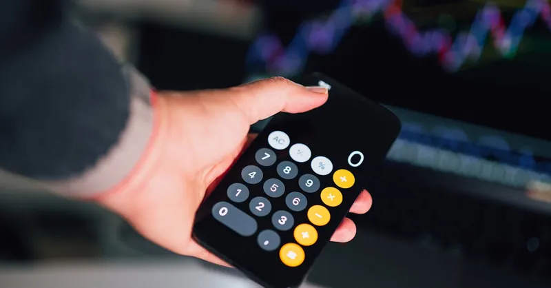 Close-up of a hand holding a smartphone calculator with financial charts on a screen in the background.