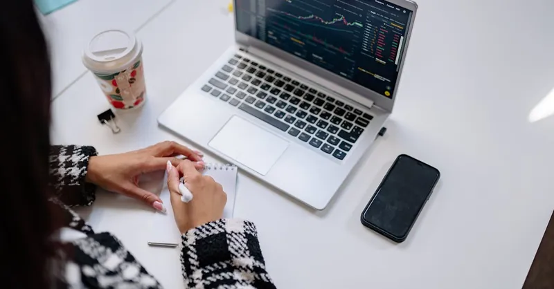 A person writing in a notebook near a laptop displaying stock charts in a modern office setting.